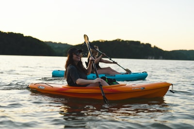 A woman wears a blue shirt and jeans on water during the day riding orange kayak
